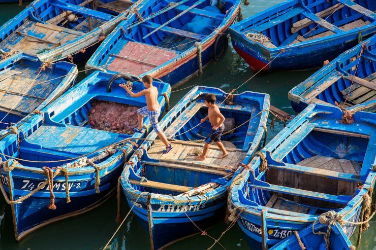 Two boys run across moored, bright blue fishing boats before jumping into the water in the Essaouira harbor.