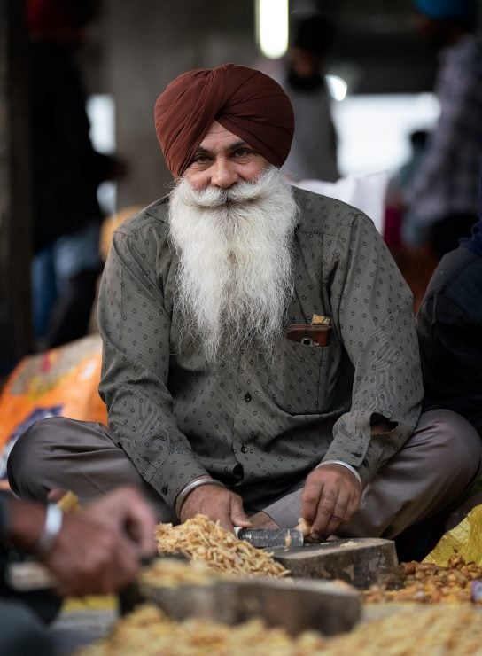 A man with a large white beard chops ginger while sitting cross legged